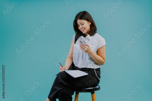 Attractive young student asian female studying while sitting on a chair and using a cellphone when doing exam isolated on light blue studio background. Learning and exam concept.