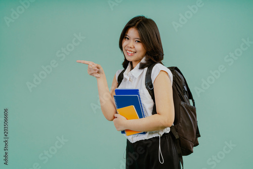 Smiling young student asian woman holding a books pointing to the side to present a product isolated on light blue studio background.