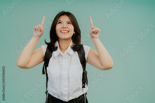 Asian college girl holding a bag and ponting of something above isolated on light green background.