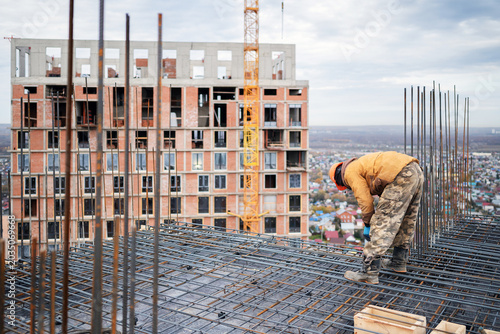 Quadro su tela An ironworker bends to tie a steel rebar grid on a high-rise concrete slab, with a brick building under construction and a yellow tower crane in the background