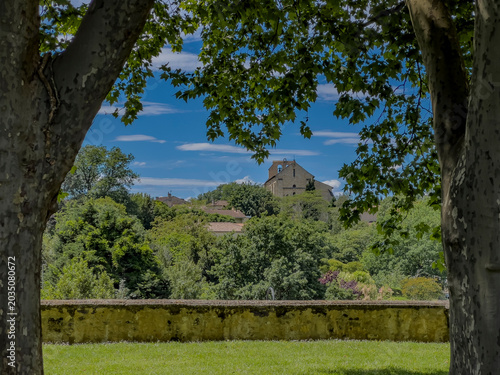 Visitors walk around the historic Abbaye de la Sauve-Majeur in Gironde, France. The ruins stand under a blue sky with clouds, revealing details of the architecture and scenery.