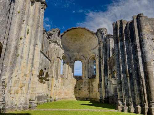 Visitors walk around the historic Abbaye de la Sauve-Majeur in Gironde, France. The ruins stand under a blue sky with clouds, revealing details of the architecture and scenery.