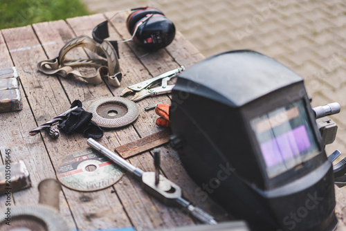 Assorted workshop tools including welding helmet and cutting discs placed on a wooden stool outdoors. Industrial DIY and metalworking equipment in natural light.