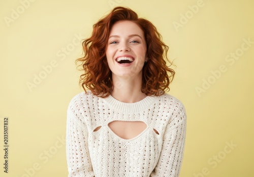 A vibrant young woman with striking red curly hair smiles brightly and laughs joyfully against a simple yellow background, exuding pure happiness and positive energy in a studio portrait