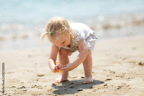 Young girl with blonde hair in a light dress crouches on a sandy beach, examining seashells while the gentle waves lap at the shore, creating a serene coastal scene. Happy childhood concept.