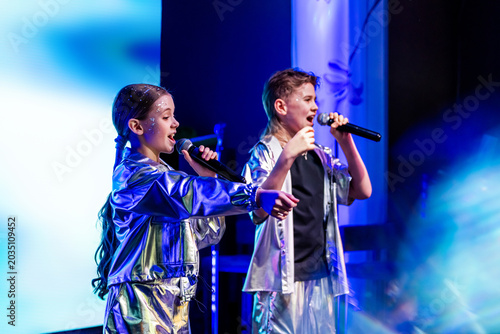 Teen girl singer performing song into microphone on stage with friend dancing. Child stage performance at music concert.