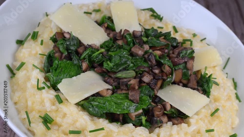A bowl contains a serving of mushroom and spinach risotto. On top are pieces of Morteau sausage and parmesan cheese. The dish is placed on a wooden surface.