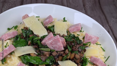 A bowl contains a serving of mushroom and spinach risotto. On top are pieces of Morteau sausage and parmesan cheese. The dish is placed on a wooden surface.