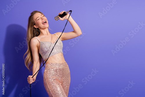 Konstfotografi Smiling young woman singing with a microphone wearing a shiny outfit against a p