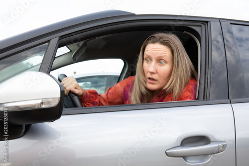 Driver woman looks surprised in car looking outside in parking lot during daytime.