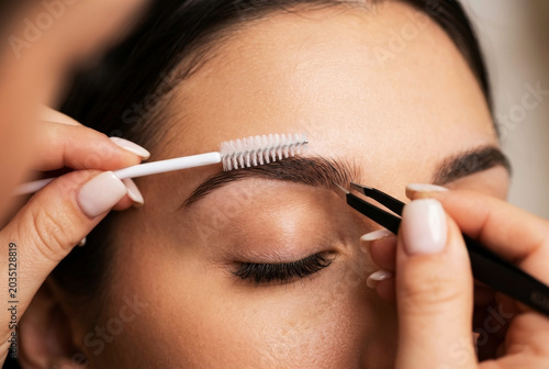 Close-up of a beauty therapist using tweezers and a spoolie brush to shape and style a woman's eyebrows during a professional brow lamination or grooming session