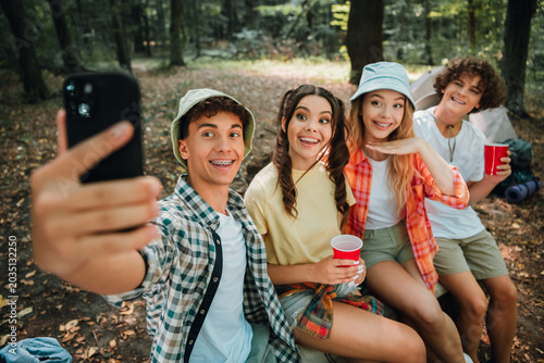 Four teenagers taking a happy group selfie while camping in the woods, smiling friends enjoying an outdoor summer hangout with cups and a tent nearby