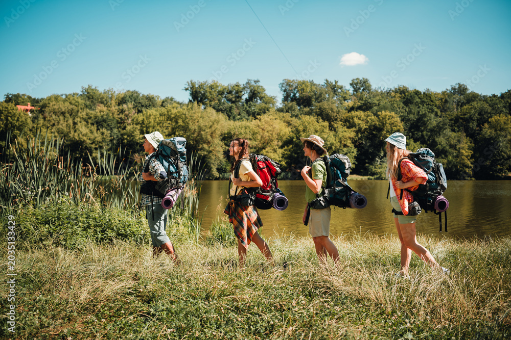 Naklejka premium Friends backpacking along a lakeside trail on a sunny summer day, hiking together with camping gear and sleeping mats in a scenic forest landscape