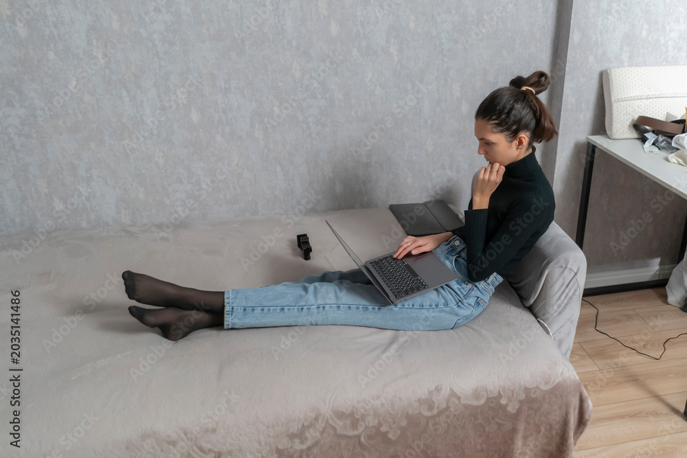 Φωτογραφία A woman sits on a bed with her legs stretched out and works on a laptop in her r