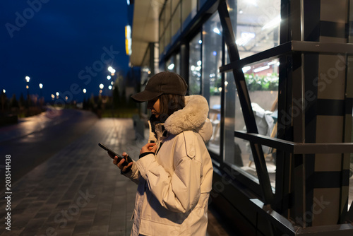Εκτύπωση καμβά Woman stands outside a building at night looking at her phone near streetlights