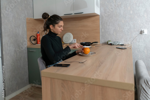 A woman is at a kitchen counter peeling an orange while sitting with a fork beside her