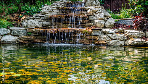 A serene pond with a small waterfall cascading down rocks into a green pond with lily pads