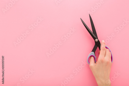 Young adult woman hand holding and showing new dark black dressmaker scissors for fabric cutting on light pink table background. Pastel color. Closeup. Empty place for text. Flat lay. Top down view.