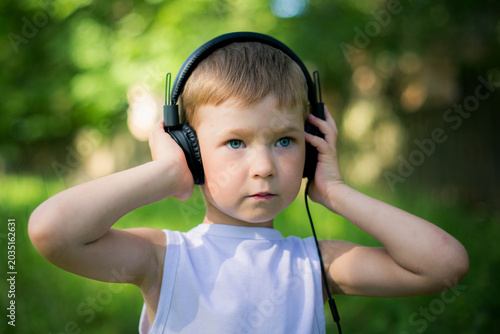 Young boy wearing headphones outdoors and listening to music in sunny green park during summer day.
