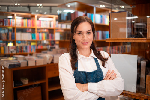 Confident Latin Brazilian Female Owner in White Shirt and Denim Apron Standing with Arms Crossed in Bookstore Medium Shot