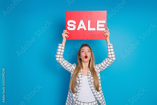 Tableau sur toile Young woman holding a red SALE sign above her head on blue background expressing