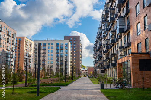 Modern residential building complex with apartment blocks and pedestrian walkway. Urban neighborhood design with landscaped courtyard, green grass and sky. Housing development for city living.