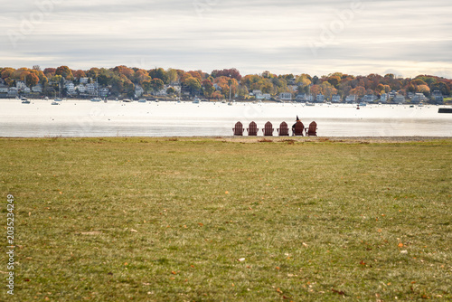 Person dressed as a witch sitting on one of a row of Adirondack chairs facing a natural harbour in Salem, MA. Houses among autum trees are in background.