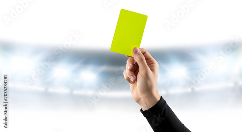 Close-up of a referee's hand holding up a bright yellow card against a blurred sports stadium background with glowing floodlights, representing a penalty or caution in a match.