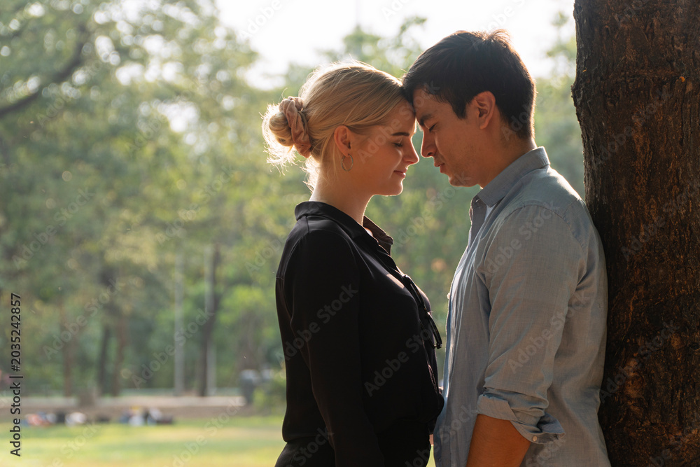 Fototapeta premium Romantic young Caucasian couple standing close with foreheads touching and eyes closed under a tree in a green park. Beautiful sunlight. Perfect for love, romance, and outdoor lifestyle concepts.