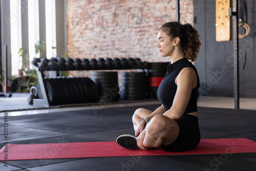 Female meditating on red mat at gym wearing black sportswear, dumbbells, rings, copy space