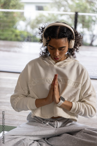 Non-binary adult meditating cross-legged on covered deck wearing cream hoodie headphones smartwatch