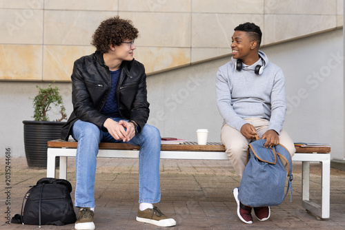 Diverse teenage males sitting on wood bench on campus holding backpacks, sharing coffee, notebook