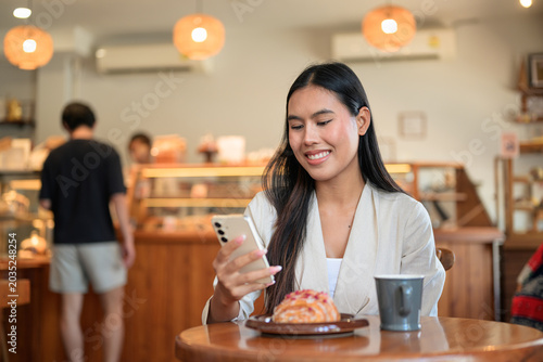 Smiling Thai businesswoman using smartphone at bakery cafe table with coffee.