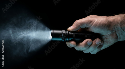 Vivid light beam from a black hand-held flashlight cutting through atmospheric smoke and shadow in a dark scene