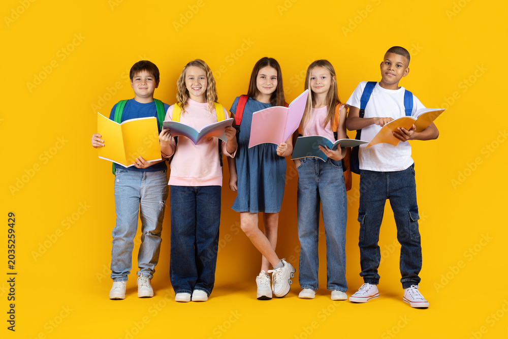 Obraz premium Happy school children with backpacks and books posing confidently on yellow studio background, representing friendship, education and modern student lifestyle