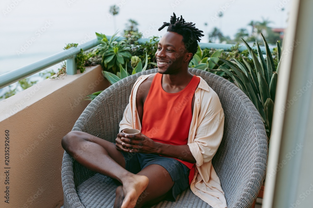 Fototapeta premium Smiling man seated in wicker balcony chair holding coffee among plants for lifestyle banner and background design