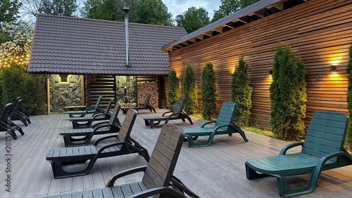 Cuadro en lienzo Cozy evening atmosphere on a wooden terrace with empty loungers, green bushes, and a rustic wall lit by warm outdoor lamps