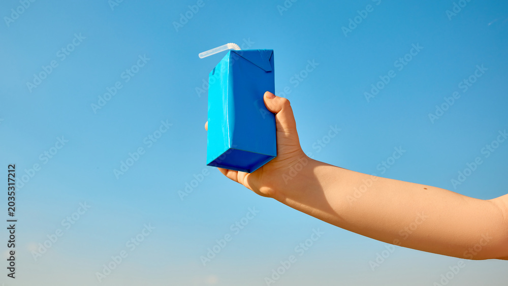 Fototapeta premium Child hand holding blue drink carton with straw against clear sky. Concept of kids beverage packaging, school lunch, dairy drink, juice box advertising, summer snack, and label mockup.