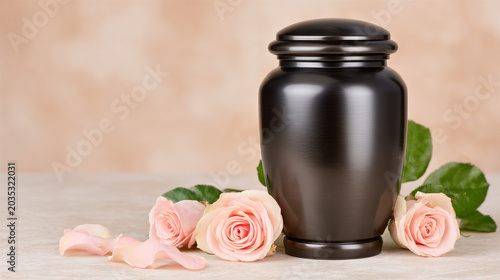 A polished dark metal cremation urn stands prominently on a light surface, accompanied by soft pink roses and scattered petals, conveying a respectful and dignified sense of remembrance.