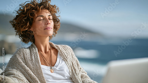 a serene woman with curly hair, eyes closed, sitting peacefully with a laptop in the foreground against a blurred ocean background.