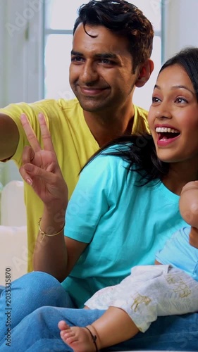 Indian family clicking smartphone selfie while sitting together on sofa in modern living room, happy parents with daughter infant baby looking at camera capturing joyful bonding moment indoors