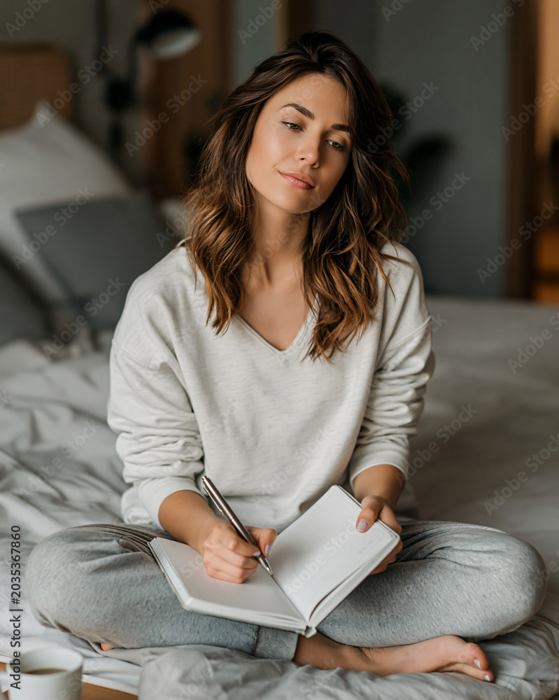 Obraz premium A Woman Writing in a Journal A thoughtful young woman sits cross-legged on a comfortable bed, holding a pen and a notebook, engrossed in writing.