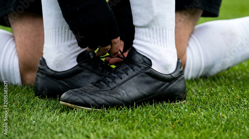 Close-up macro shot of a football player bending down on the pitch to tie the laces of plain black leather boots.