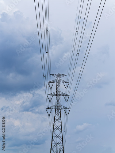 High-voltage power lines stretch against the sky. Steel towers rise strong in the open field. Clouds swirl with tension and power. The scene hints at modern energy's reach