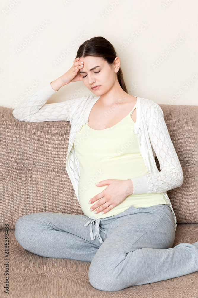 Obraz premium Young pregnant woman with headache sitting on sofa. Pregnancy expectation concept