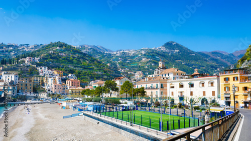 Minori Italy coastal town with hillside terraces, colorful buildings, beach and sports field under clear blue sky. Minori architecture blends with green hills creating scenic Mediterranean landscape
