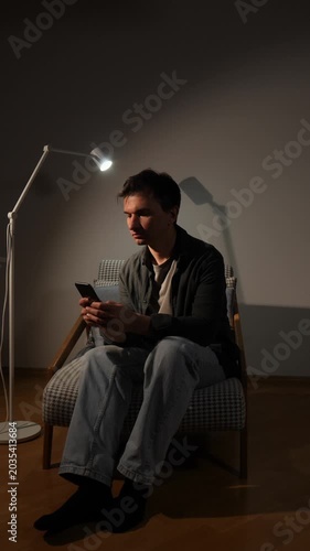 Vertical video man focusing on his smartphone screen in a dimly lit room, depicting concepts of loneliness, social isolation, technology addiction, and silent preoccupation