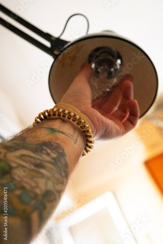 A man's hand screws the bulb of a desk lamp. The man's forearm is tattooed. The image recalls several concepts including do-it-yourself and electricity