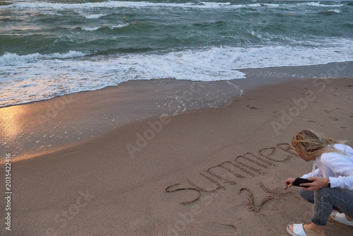 Blonde female blogger writing on a sandy beach at sunset, sea waves on background.