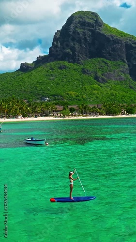 Aerial view of woman in white swimsuite standing on sup board with paddle on crystal turquoise water with coral reef on the tropical island in Indian ocean. Active water sport. Mauritius island